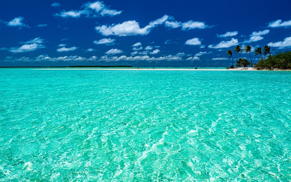 Mare turchese con spiaggia e palme in lontananza cielo azzurro e nuvolette bianche